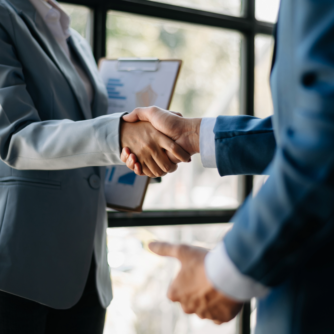 Two professionals shake hands in a bright office, one holding a clipboard with charts and graphs in view.