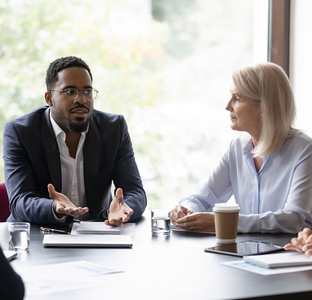 Two professionals in a business meeting at a conference table, talking with notebooks, papers, and coffee nearby.