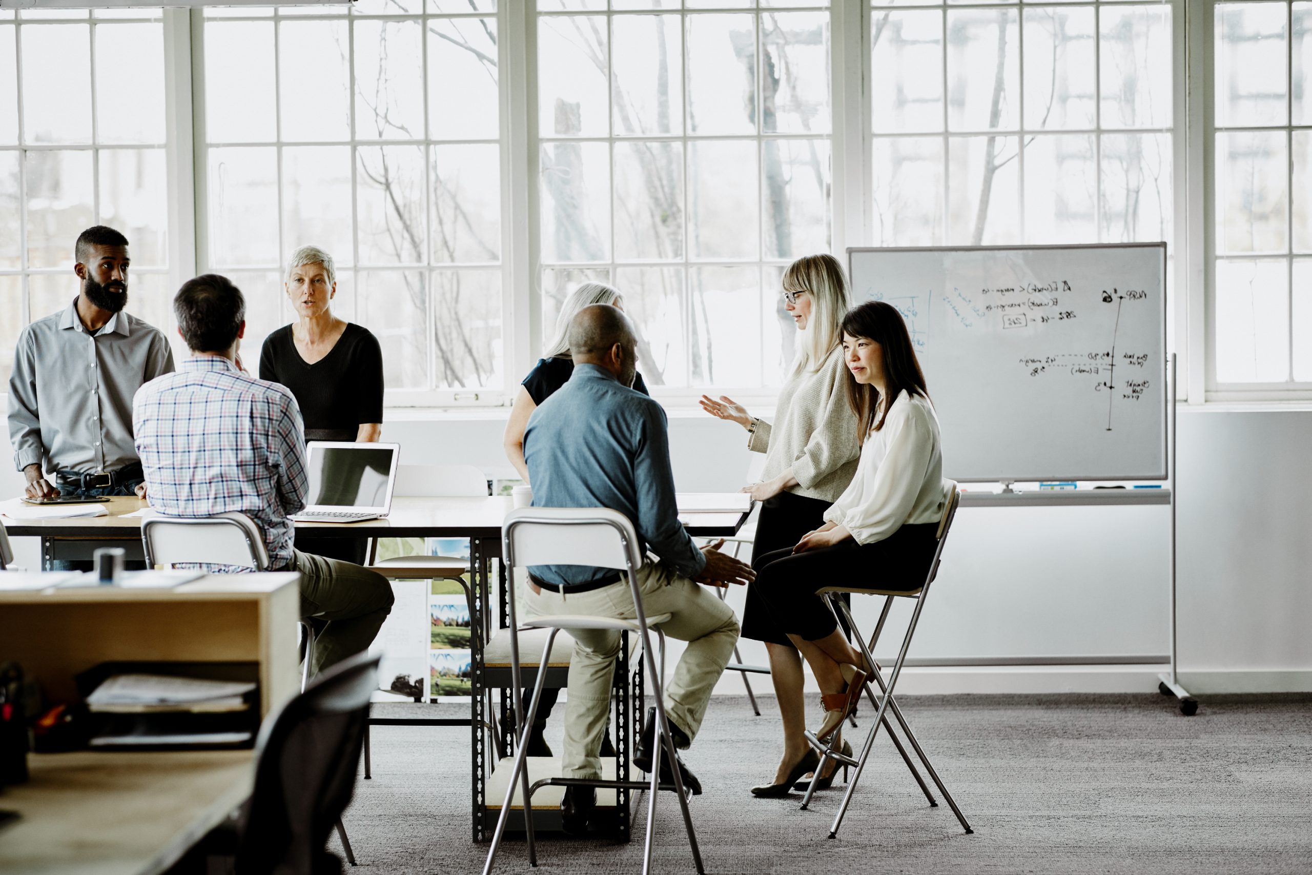 A diverse group of professionals standing and sitting around a conference table, discussing near a whiteboard in a bright room with large windows.