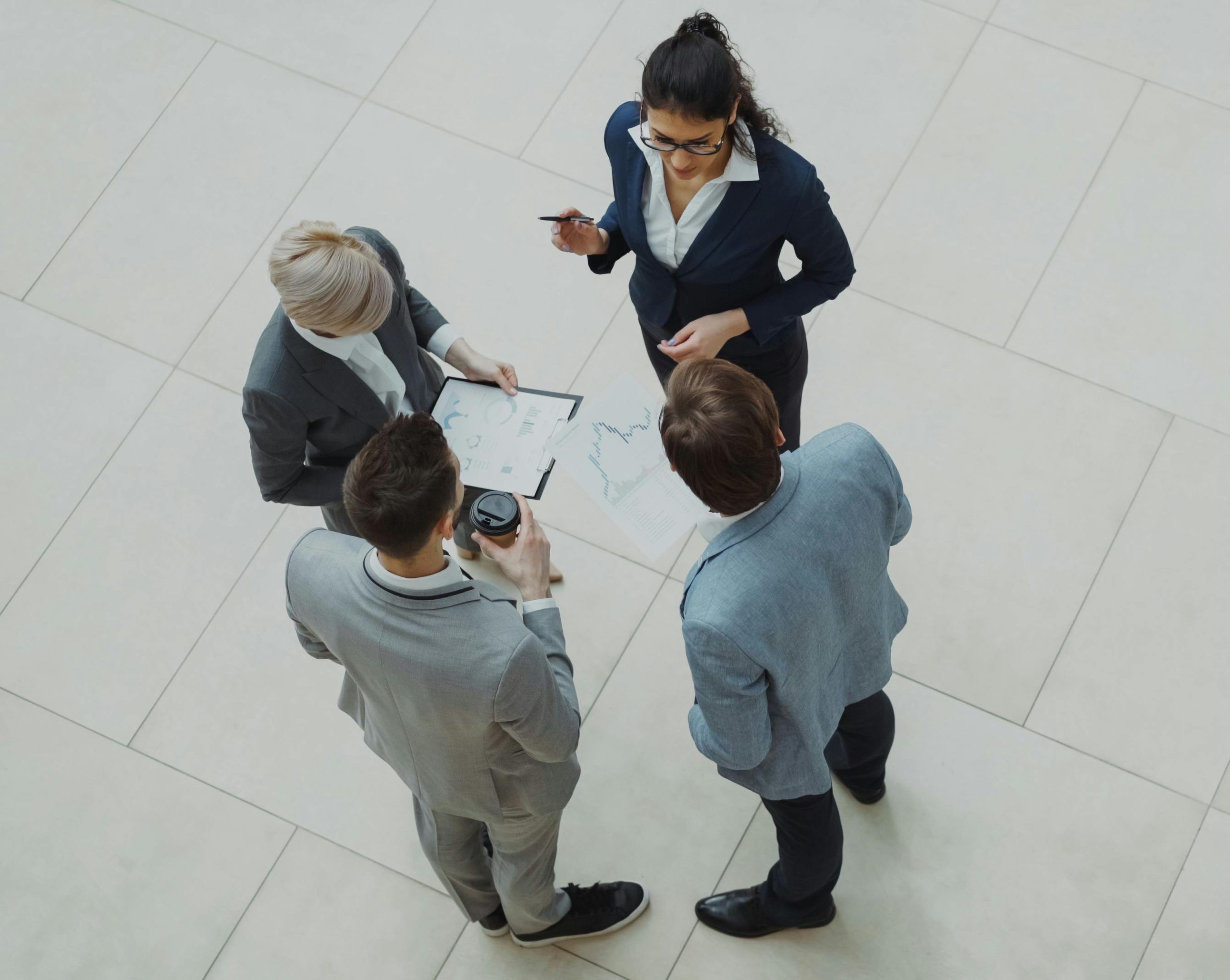 Overhead view of five professionals in business attire standing in a circle, reviewing charts and documents on the tiled floor