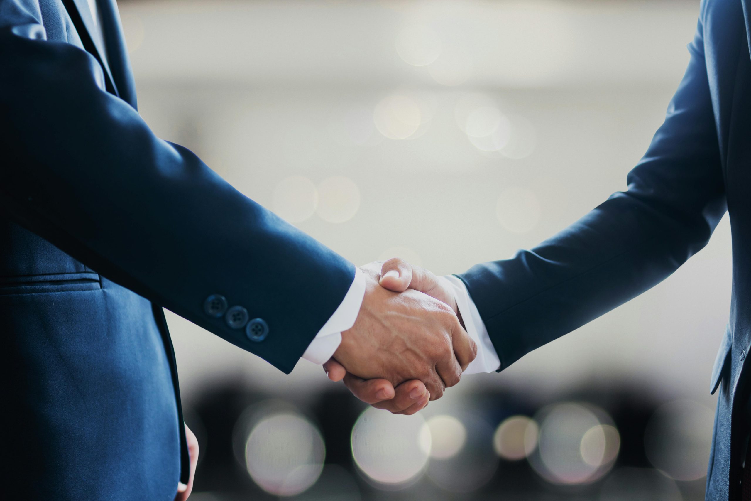 Two businesspeople in dark suits shake hands in a professional setting.